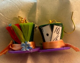 Puede incluir: Un par de pendientes caprichosos estilo Sombrerero Loco. Cada sombrero en miniatura presenta un ala morada, una cinta naranja y una parte superior verde adornada con plumas de colores o naipes. Los ganchos dorados completan el diseño.