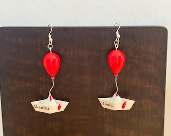 Puede incluir: Un par de pendientes colgantes con un globo rojo brillante unido a un barco de papel blanco. El barco tiene el texto SS Georgie impreso en el lateral en letras negras, con pequeños detalles rojos en la parte superior de la estructura.