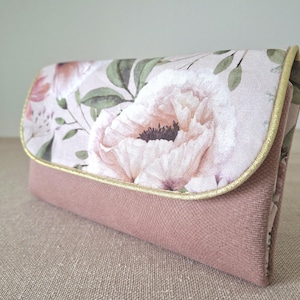May include: A pink clutch bag with a floral design on the flap. The flap displays a large, pale pink flower with green leaves. The base of the bag is solid pink, and the edges are trimmed with gold.