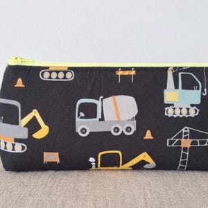 Puede incluir: Estuche negro con cremallera amarilla, con un diseño de vehículos de construcción. El diseño incluye ilustraciones de excavadoras, hormigoneras y grúas en tonos grises, amarillos y azules. Perfecto para guardar lápices.
