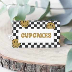 以下が含まれることがあります： 黒と白の市松模様の看板に、金のスマイリーフェイスと金の文字で「CUPCAKES」と書かれている。