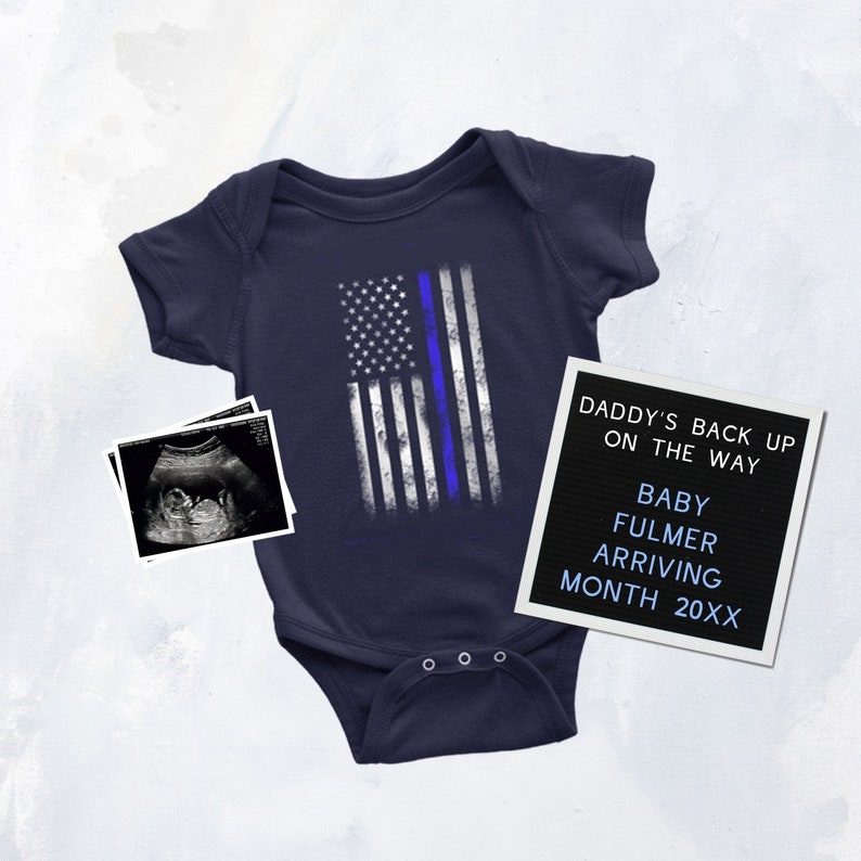 Puede incluir: Un body de beb&eacute; azul marino con un dise&ntilde;o de bandera estadounidense blanca y azul con una fina l&iacute;nea azul. Un tablero de letras negro con el texto "Daddy's back up on the way Baby Fulmer arriving month 20XX". Una imagen de ultrasonido en blanco y negro est&aacute; en el fondo.