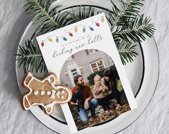 May include: Christmas card featuring a family of three standing in front of a house, with the text "This Christmas we are decking new halls" and a string of colorful lights above the text.