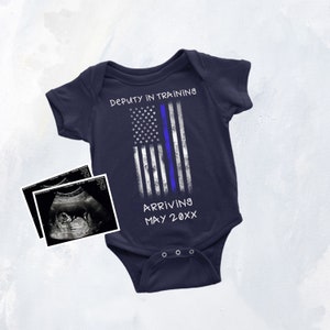 Puede incluir: Un body de beb&eacute; azul marino con un dise&ntilde;o de bandera estadounidense blanca y azul. El texto "Deputy in Training" y "Arriving May 20XX" est&aacute; impreso en el body. Una imagen de ultrasonido en blanco y negro est&aacute; en el fondo.