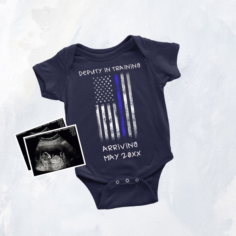 Puede incluir: Un body de beb&eacute; azul marino con un dise&ntilde;o de bandera estadounidense blanca y azul. El texto "Deputy in Training" y "Arriving May 20XX" est&aacute; impreso en el body. Una imagen de ultrasonido en blanco y negro est&aacute; en el fondo.