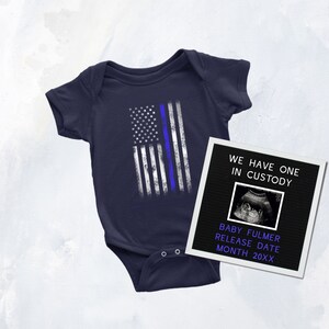 Puede incluir: Un body de beb&eacute; azul marino con un dise&ntilde;o de bandera estadounidense desgastado con una l&iacute;nea azul. Un marco de fotos en blanco y negro con el texto "We have one in custody" y una imagen de ecograf&iacute;a est&aacute; al lado del body. El texto "Baby Fulmer Release Date Month 20XX" est&aacute; debajo de la imagen de la ecograf&iacute;a.