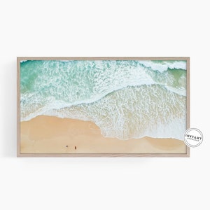 May include: An aerial view of a sandy beach with turquoise water and white foamy waves crashing on the shore. Two people are walking on the beach.