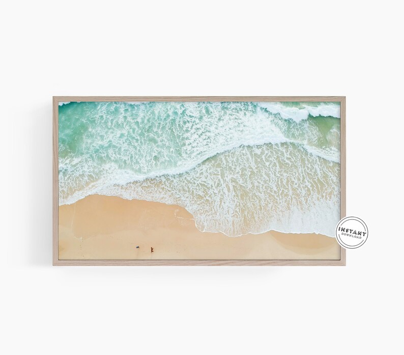 May include: An aerial view of a sandy beach with turquoise water and white foamy waves crashing on the shore. Two people are walking on the beach.