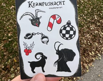 May include: A rectangular sticker with the text "Krampusnacht" and various black and white illustrations. The illustrations include a goat head, candy cane, handcuffs, holly berries, a checkered ball, a goat, and a Krampus figure.