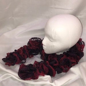 May include: A burgundy and black crocheted scarf with a ruffled texture. The scarf is displayed on a white mannequin head.