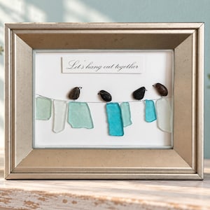 May include: A framed wall art piece featuring four dark pebble birds perched on a wire above several pieces of light blue and teal sea glass hanging like laundry. The white background includes the printed text Let's hang out together.