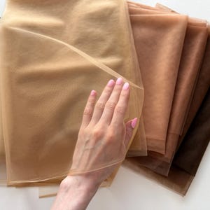 May include: A collection of sheer, fine-mesh fabric swatches arranged in a fan shape. The fabric pieces display a gradient of skin-tone shades, ranging from light beige and tan to medium brown and deep espresso, featuring a soft, translucent texture.