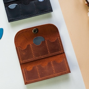 May include: Three leather guitar pick holders in different colors: brown, black, and orange. Each holder has multiple slots for storing guitar picks.