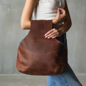 May include: A brown leather tote bag with a circular monogram "ST" on the front. The bag has a wide, open top and a single strap.