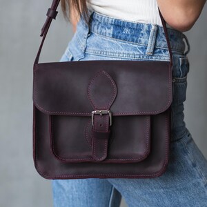 Puede incluir: Bolso bandolera de cuero morado oscuro con cierre de solapa y bolsillo delantero. El bolso presenta una hebilla y correa plateadas, con costuras visibles en un color morado más claro. El bolso se lleva al hombro.