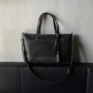 Puede incluir: Un bolso tote de cuero negro con una correa larga y un detalle de borla. El bolso tiene un compartimento grande y un bolsillo m&aacute;s peque&ntilde;o en la parte delantera.