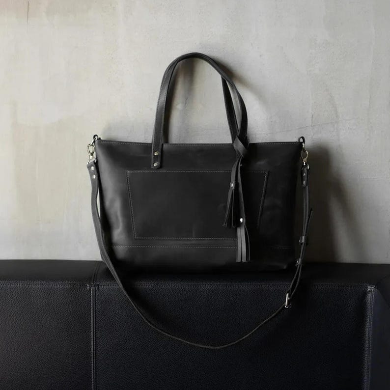 Puede incluir: Un bolso tote de cuero negro con una correa larga y un detalle de borla. El bolso tiene un compartimento grande y un bolsillo m&aacute;s peque&ntilde;o en la parte delantera.