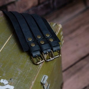 May include: Black leather wrist cuffs with brass buckles and rivets. The cuffs are arranged on a weathered, olive-green wooden surface, with a blurred brick background. The leather appears to be thick and durable.
