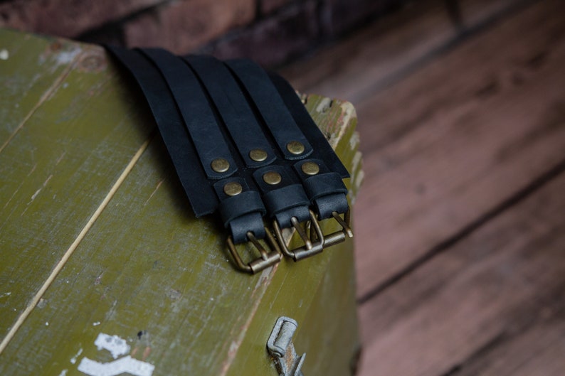 May include: Black leather wrist cuffs with brass buckles and rivets. The cuffs are arranged on a weathered, olive-green wooden surface, with a blurred brick background. The leather appears to be thick and durable.