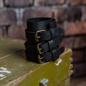 May include: A stack of black leather wrist cuffs, each secured with a brass buckle. The cuffs are made from thick leather and are designed to be worn around the wrist. The buckles have a vintage appearance. They are stacked on a green wooden box.