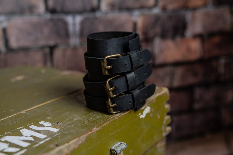 May include: A stack of black leather wrist cuffs, each secured with a brass buckle. The cuffs are made from thick leather and are designed to be worn around the wrist. The buckles have a vintage appearance. They are stacked on a green wooden box.