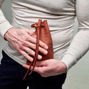 Pu&ograve; includere: Una borsa in pelle marrone con chiusura a coulisse. La borsa &egrave; tenuta nelle mani di una persona.