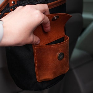 Op de afbeelding: Een zwarte canvas tas met een bruin leren zakje en een magnetische sluiting. Het zakje is open en de tas hangt aan een autostoel.