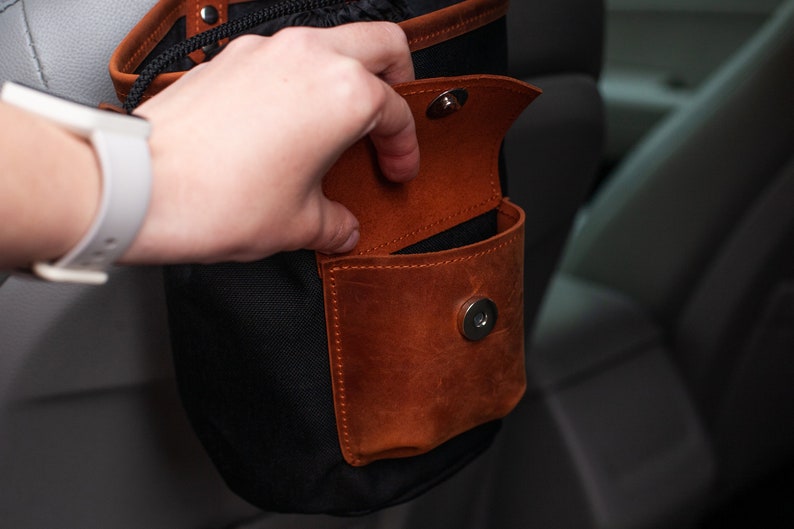 Op de afbeelding: Een zwarte canvas tas met een bruin leren zakje en een magnetische sluiting. Het zakje is open en de tas hangt aan een autostoel.