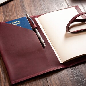 May include: A burgundy leather journal with a pen holder and a passport pocket. The journal has a strap closure and is open to reveal blank pages.