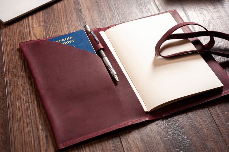 May include: A burgundy leather journal with a pen holder and a passport pocket. The journal has a strap closure and is open to reveal blank pages.