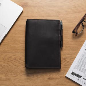 May include: A dark brown leather notebook with a pen tucked into the cover. The notebook is on a wooden surface with a laptop and a pair of glasses in the background.
