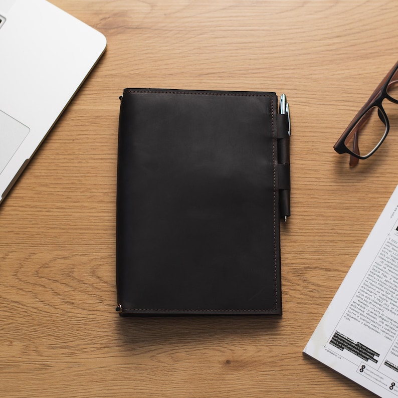 May include: A dark brown leather notebook with a pen tucked into the cover. The notebook is on a wooden surface with a laptop and a pair of glasses in the background.