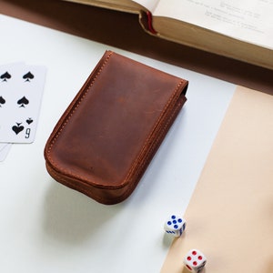 May include: A brown leather case for playing cards, dice, or other games. The case is closed and sitting on a white surface with playing cards and dice nearby.