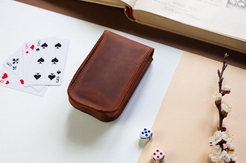 May include: A brown leather case for playing cards, dice, or other games. The case is closed and sitting on a white surface with playing cards and dice nearby.