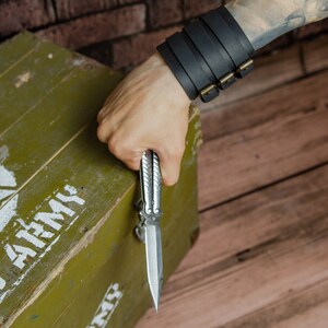 May include: A silver butterfly knife is held in a hand, with a black leather cuff bracelet with brass buckles on the wrist. The knife is open, revealing its sharp blade. An olive green wooden box with a white star and "U.S. ARMY" text is in the background.