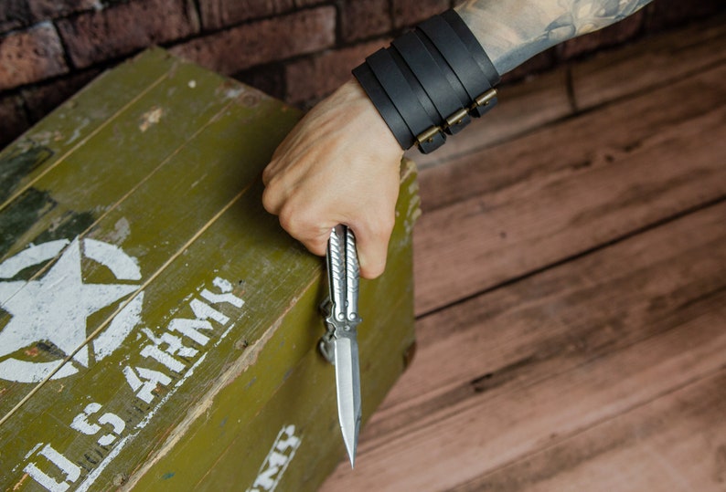 May include: A silver butterfly knife is held in a hand, with a black leather cuff bracelet with brass buckles on the wrist. The knife is open, revealing its sharp blade. An olive green wooden box with a white star and "U.S. ARMY" text is in the background.