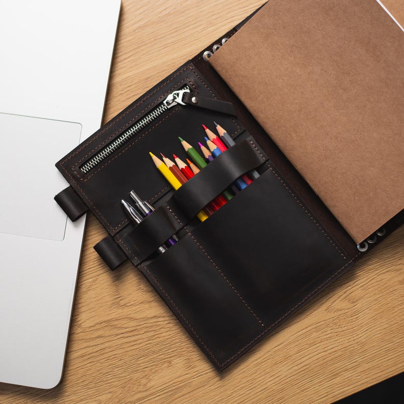 May include: A brown leather notebook with a zipper closure and multiple pockets for pens and pencils. The notebook is open and has a blank page inside.