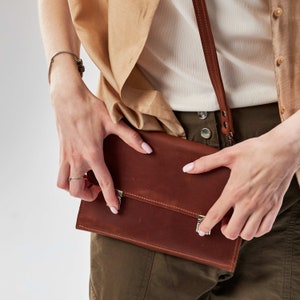 Op de afbeelding: Een bruine leren clutch met twee zilveren sluitingen. De clutch wordt vastgehouden door een persoon die een beige jas en een wit shirt draagt.