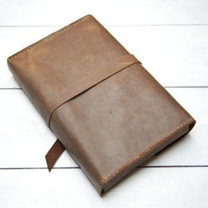 May include: A brown leather journal with a strap closure. The journal is closed and lying on a white wooden surface.