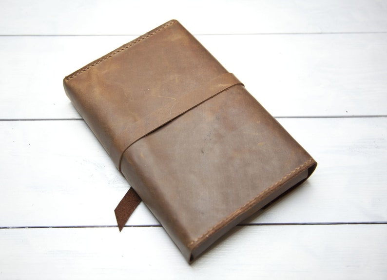 May include: A brown leather journal with a strap closure. The journal is closed and lying on a white wooden surface.
