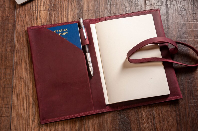 May include: A burgundy leather journal with a pen holder and a passport pocket. The journal has a blank page open and a leather strap.