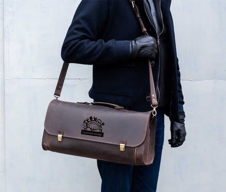 Pu&ograve; includere: Una borsa a tracolla in pelle marrone con un design inciso personalizzato che raffigura una mano che tiene degli strumenti. La borsa ha una lunga tracolla regolabile e una chiusura a lembo con due fibbie in ottone.