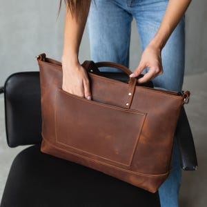 Puede incluir: Un bolso tote de cuero marr&oacute;n con cierre de cremallera y un bolsillo frontal grande. El bolso tiene un asa superior y una correa para el hombro.