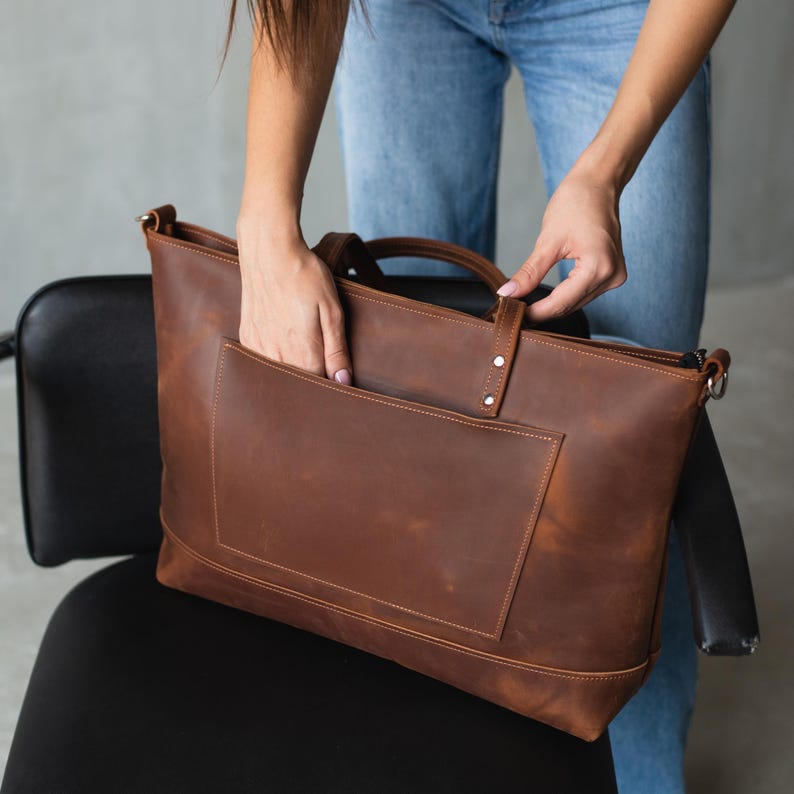 Puede incluir: Un bolso tote de cuero marr&oacute;n con cierre de cremallera y un bolsillo frontal grande. El bolso tiene un asa superior y una correa para el hombro.