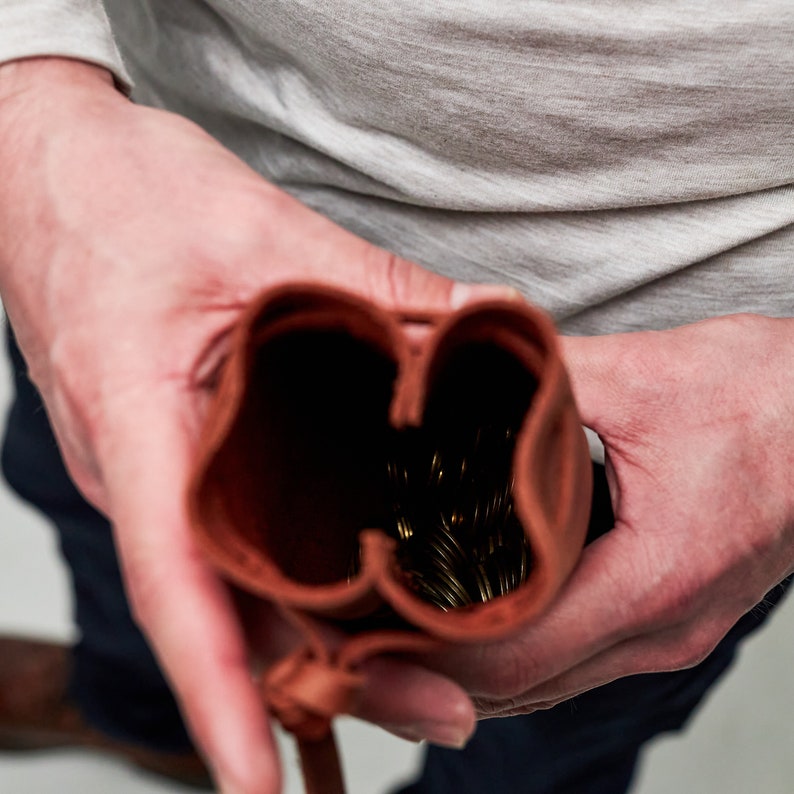 Pu&ograve; includere: Una borsa in pelle marrone con chiusura a cordoncino, aperta per rivelare un mucchio di monete dorate all'interno.