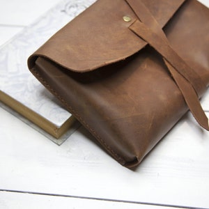 May include: A brown leather clutch with a flap closure and a long strap. The strap is tied in a knot and has a metal button.