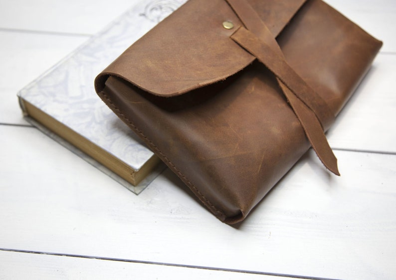 May include: A brown leather clutch with a flap closure and a long strap. The strap is tied in a knot and has a metal button.