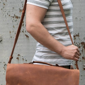 Pu&ograve; includere: Una borsa a tracolla in pelle marrone con una lunga tracolla regolabile. La borsa ha due fibbie metalliche e una cerniera sul davanti.