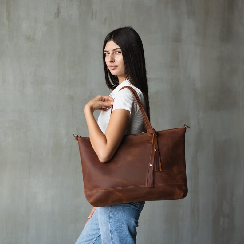 Puede incluir: Un bolso tote de cuero marr&oacute;n con un borla que cuelga de la cremallera. El bolso lo lleva una persona que lleva una camisa blanca y unos vaqueros azules.