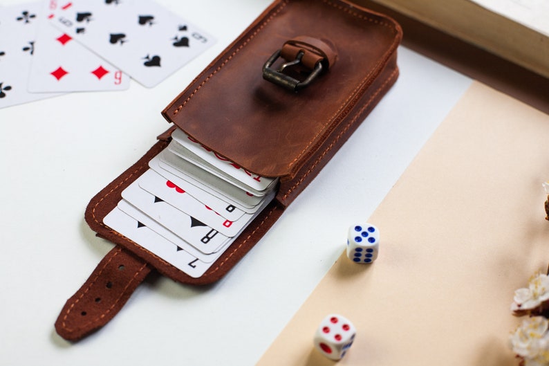 May include: A brown leather card case with a buckle closure. The case is open and a deck of playing cards is visible inside. Two white dice with blue dots are on a beige surface.